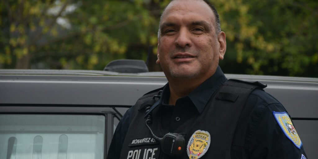 Officer Edwin Monarrez, in uniform, stands for a portrait in front of his police vehicle while on shift at the Laurel Police Department early in the morning during an overcast day.