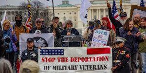 15 veteran protestors stand in front of the U.S. Capitol building. A large banner reads "Stop the deportation of military veterans," and another sign reads "Support our veterans." American and veteran flags are visible.